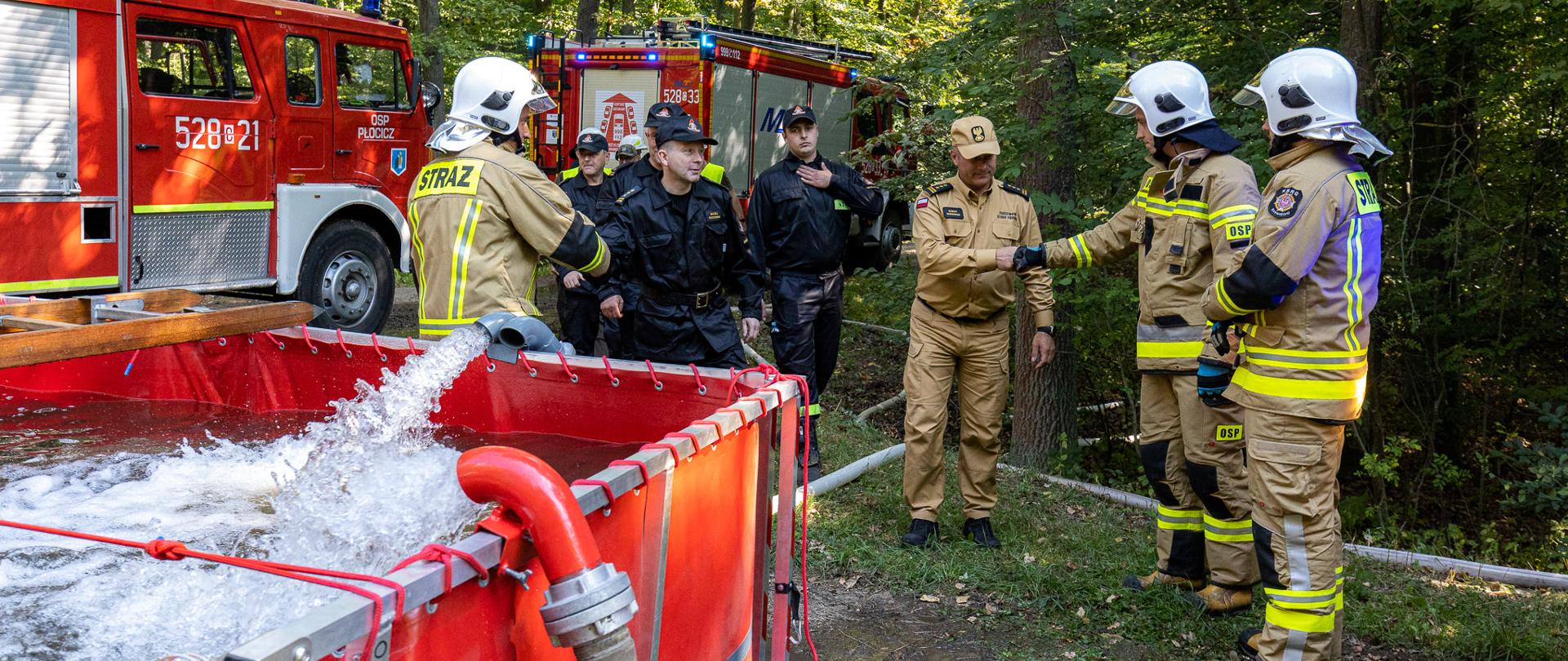 Zdjęcie przedstawia strażaków w lesie przy zbiorniku wodnym. Jedne ze strażaków wita się z zastępcą komendanta wojewódzkiego PSP w tle samochody pożarnicze
