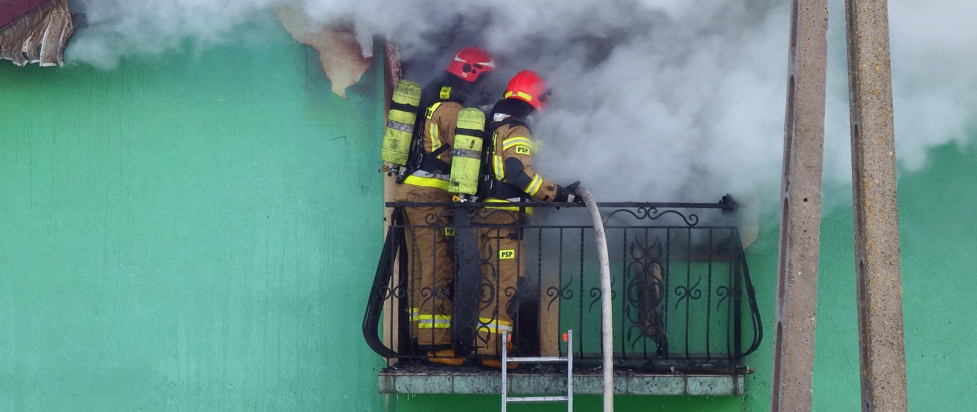 Zdjęcie przedstawia dwóch strażaków stojących na balkonie na tle zielonej ściany domu. Ubrani są w obrania bojowe, na plecach mają aparaty powietrzne a na głowach czerwone hełmy. Z okna wydobywają się kłęby dymu spowijające strażaków. Do balkonu przystawiona jest drabina, a przez balustradę przełożony jest napełniony wodą wąż strażacki.