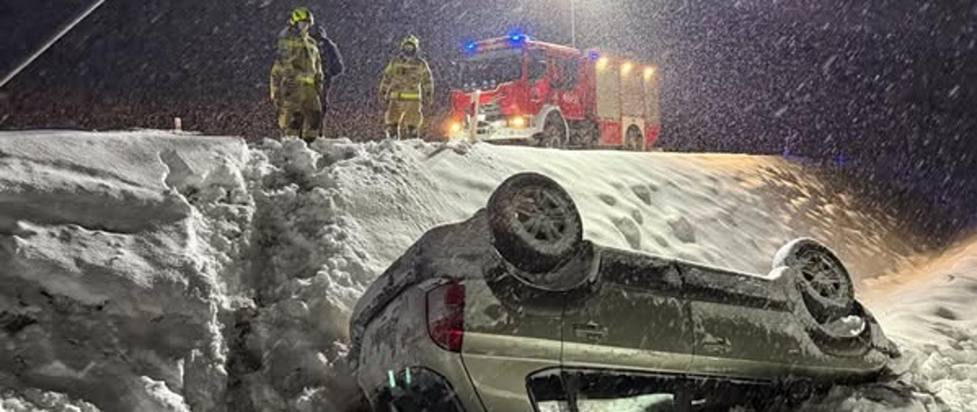 Zdjęcie zrobione nocą. Samochód osobowy leży na dachu w głębokim rowie, dużo śniegu. Wyżej na drodze Samochód strażacki i strażacy. Pada śnieg.