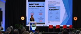 Na zdjęciu widać publiczność tyłem, która spogląda na ekran oraz prelegentkę. Prelegentka stoi przy mównicy, za nią znajduje się ekran, na którym widnieją informacje o nutriach w Rybniku.