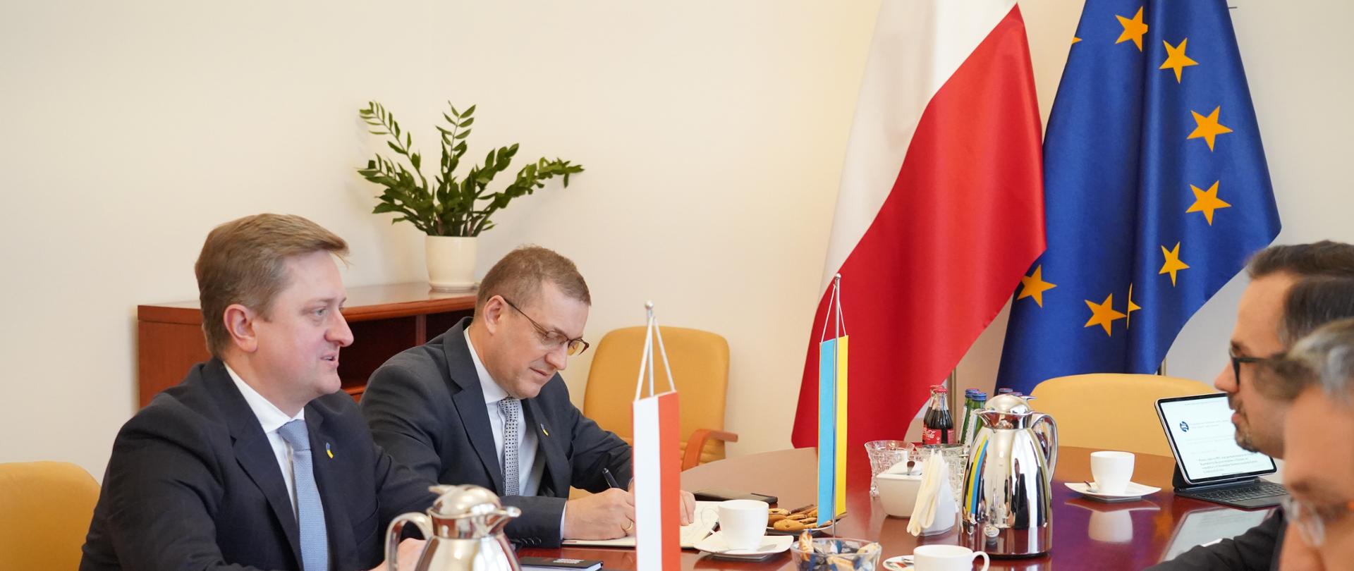 Cztery osoby siedzą przy stole. Dwie z lewej i dwie z prawej strony.