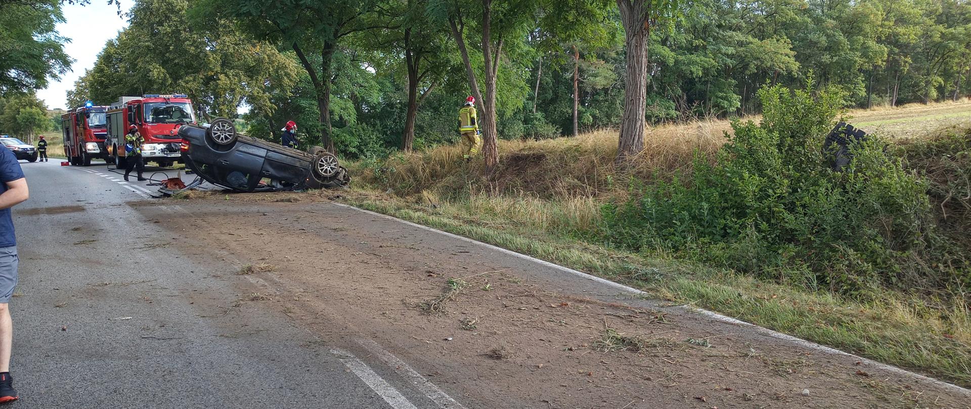 Samochód leży na dachu na pasie drogi rozsypana jest ziemia z pobocza drogi. W oddali stoją samochody PSP.