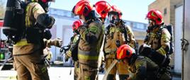 Kilku strażaków stoi w grupie, ubrani są w piaskowy ubranie specjalne, niektórzy na plecach mają założone aparaty ochrony dróg oddechowych. Jeden z nich przykucnął. Na głowach mają czerwone kaski. W tle budynki zakładów.