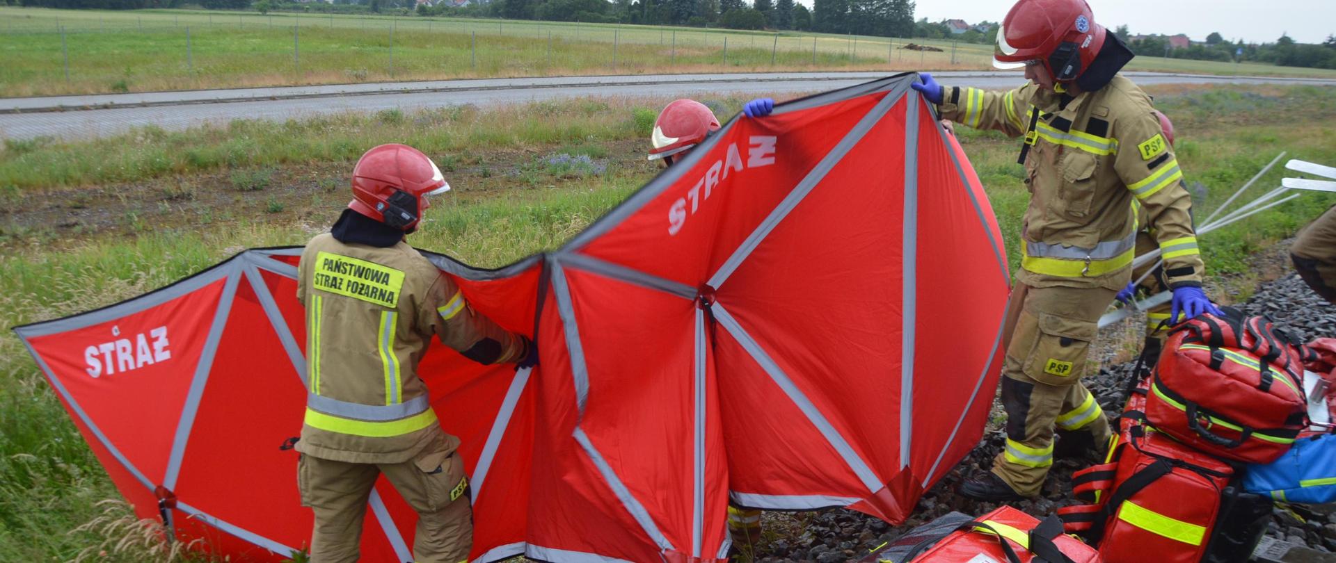 Wypadek na torach ze skutkiem śmiertelnym Szlak kolejowy Rawicz-Wrocław. Z lewej strony torowiska 4 strażaków ustawia czerwony parawan zabezpieczający. Z prawej strony znajduje sie sprzęt medyczny. W tle łąki i drzewa. 