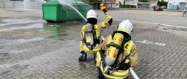dwóch strażaków w ubraniach specjalnych ubrany w aparaty powietrzne podaje prąd strumień wody na zielony duży kontener na śmieci jeden z nich podnosi rękę z sygnałem gotowości, drugi za nim podtrzymuje wąż