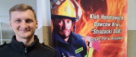 Kolorowa fotografia wykonana w pomieszczeniu. Na pierwszym planie przedstawia strażaka trzymającego legitymację honorowego dawcy krwi. Strażak stoi na tle plakatu Honorowych Dawców Krwi „Strażacki Dar” przy Komendzie Powiatowej PSP w Brzozowie.