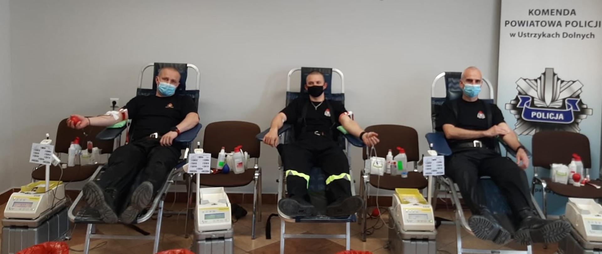 Na zdjęciu widać 3 funkcjonariuszy KP PSP w Ustrzykach Dolnych siedzących na fotelach w momencie oddawania krwi. Za nimi godło Polski oraz logo i napis "Policja Komenda Powiatowa Policji w Ustrzykach Dolnych. Obok nich krzesła, maszyny do pobierania krwi, różne medykamenty oraz kosze na odpady.