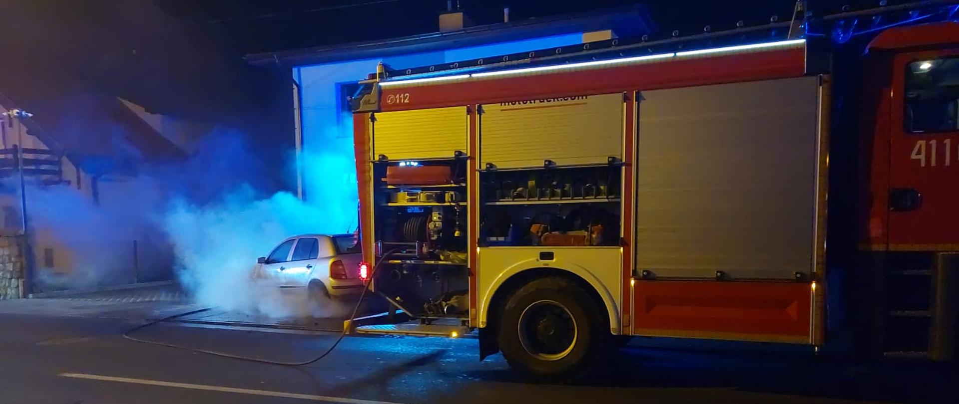 Pora nocna, zdjęcie na pierwszym palnie widać tył czerwonego samochodu straży pożarnej zaparkowanego na asfaltowej drodze. Samochód ma otwarte rolety zabudowy służącej do przewozu sprzętu. Za samochodem straży pożarnej widać srebrny samochód osobowy, z którego intensywnie wydobywa się szary dym. Za samochodem widać budynki. 