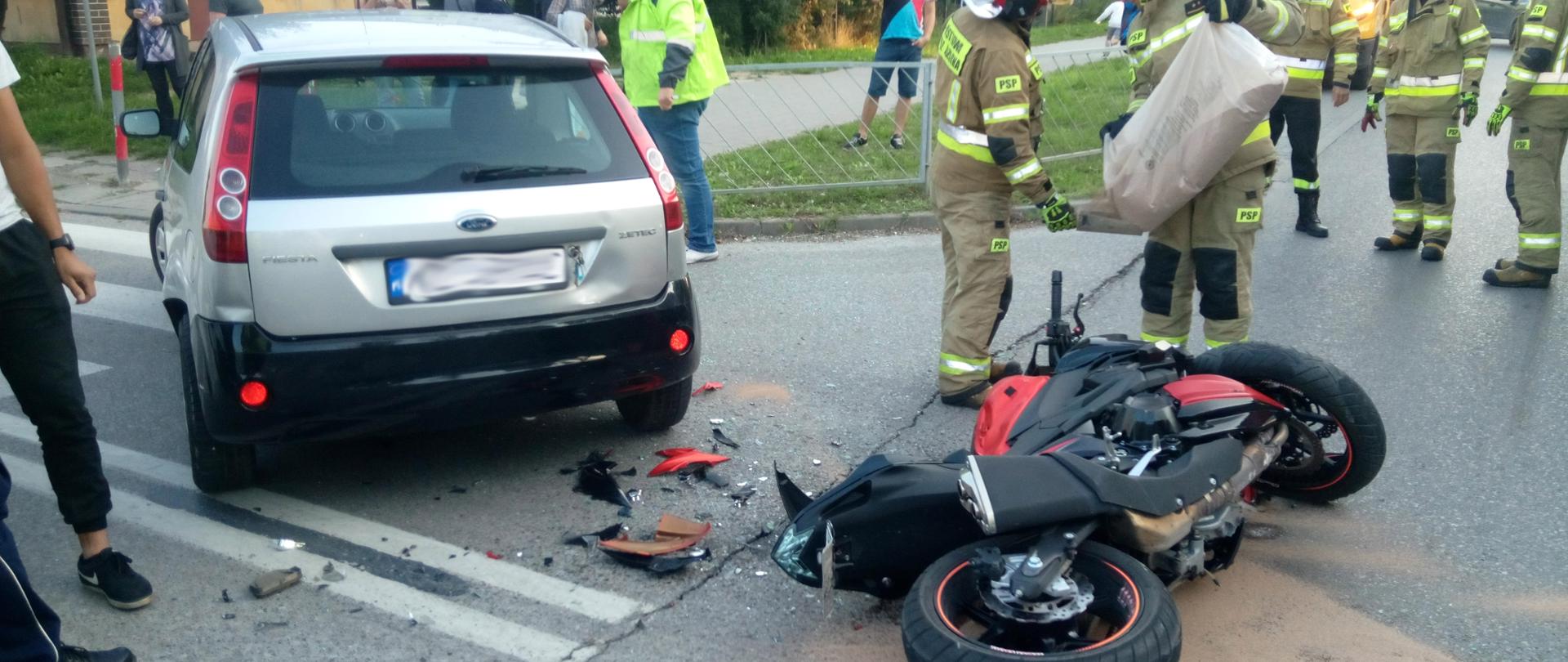 Zdjęcie przeszkadza przewrócony motocykl Kawasaki oraz samochód osobowy stojący tyłem - ford fusion. Za pojazdami strażacy neutralizują plamę za pomocy sorbentu. w tle widać świadków zdarzenia oraz pozostałych strażaków.