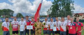 Grupa polskich strażaków w strojach sportowych(granatowe spodenki i biało czerwone koszulki) na zdjęciu grupowym, po środku grupy stoi strażak PSP w mundurze służbowym, za nim powiewa flaga RP