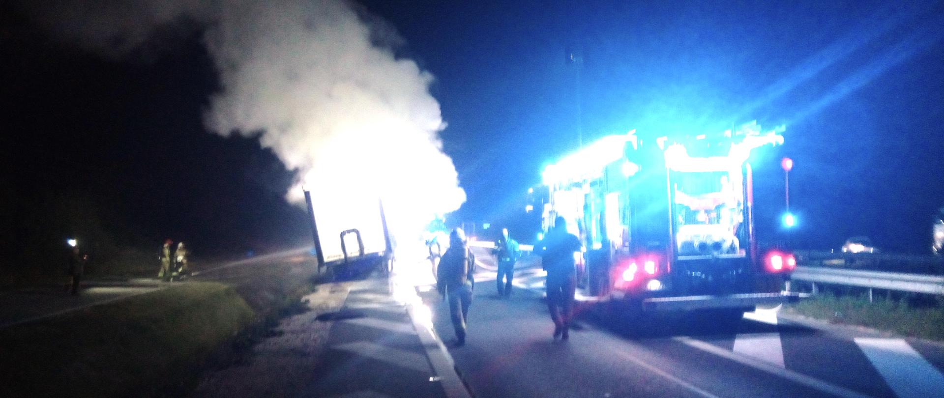 Zdjęcie przedstawia ulicę na której doszło do pożaru naczepy ciągnika siodłowego. Na drodze stoi wóz strażacki, przemieszczają się funkcjonariusze służb ratowniczych a na poboczu po lewej stronie stoi naczepa, z której unoszą się kłęby dymu i pary wodnej.