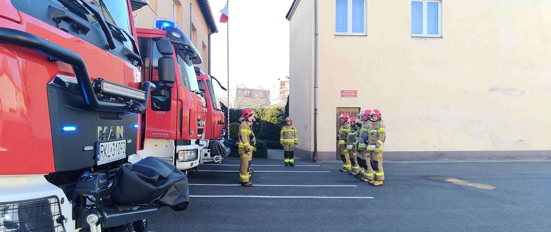 Na asfaltowym placu w dwóch szeregach oddalonych od siebie kilka metrów stoją strażacy w umundurowaniu w kolorze piaskowym. Po lewej stronie zdjęcia widać przody trzech samochodów strażackich. W tle budynki oraz pomiędzy nimi masz z zawieszoną flagą.