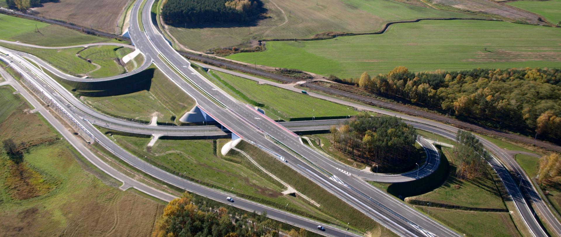 Zdjęcie przedstawiające węzeł drogowy Poznań Północ na drodze ekspresowej S11. Węzeł widoczny z powietrza przebiegający między łąkami, polami i zabudową magazynową. Na jednej z łącznic doprowadzających ruch do węzła widoczne poruszające się pojazdy.