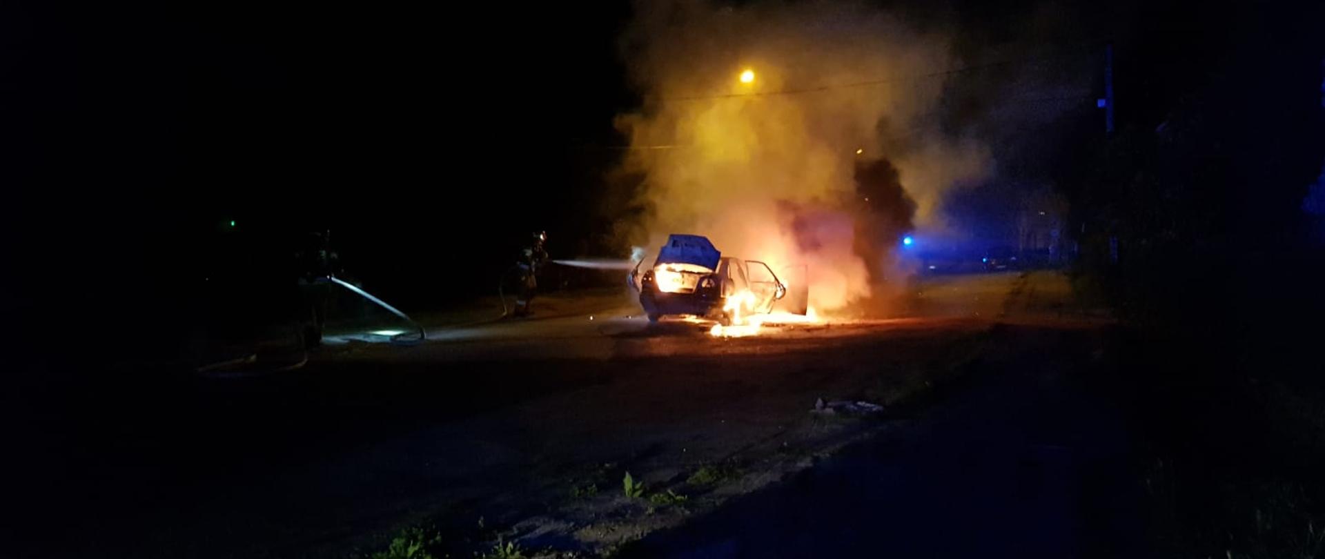 Pora nocna na zdjęciu centralnie pali się samochód osobowy, a obok strażak prowadzi akcję gaśniczą. samochód jest cały objęty ogniem, występuje duże zadymienie.