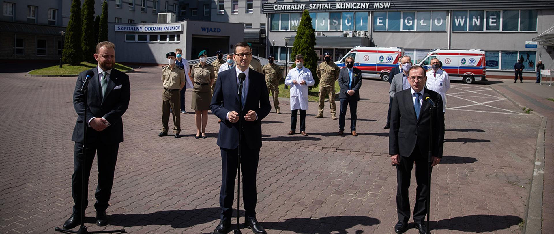 Na pierwszym planie stoją Minister Zdrowia Łukasz Szumowski, Premier Mateusz Morawiecki, Minister MSWiA Mariusz Kamiński. W tle CSK MSWiA, stojące dwie karetki, stoją również m. in. lekarze, żołnierze i policjanci zaangażowani w walkę z pandemią koronawirusa.