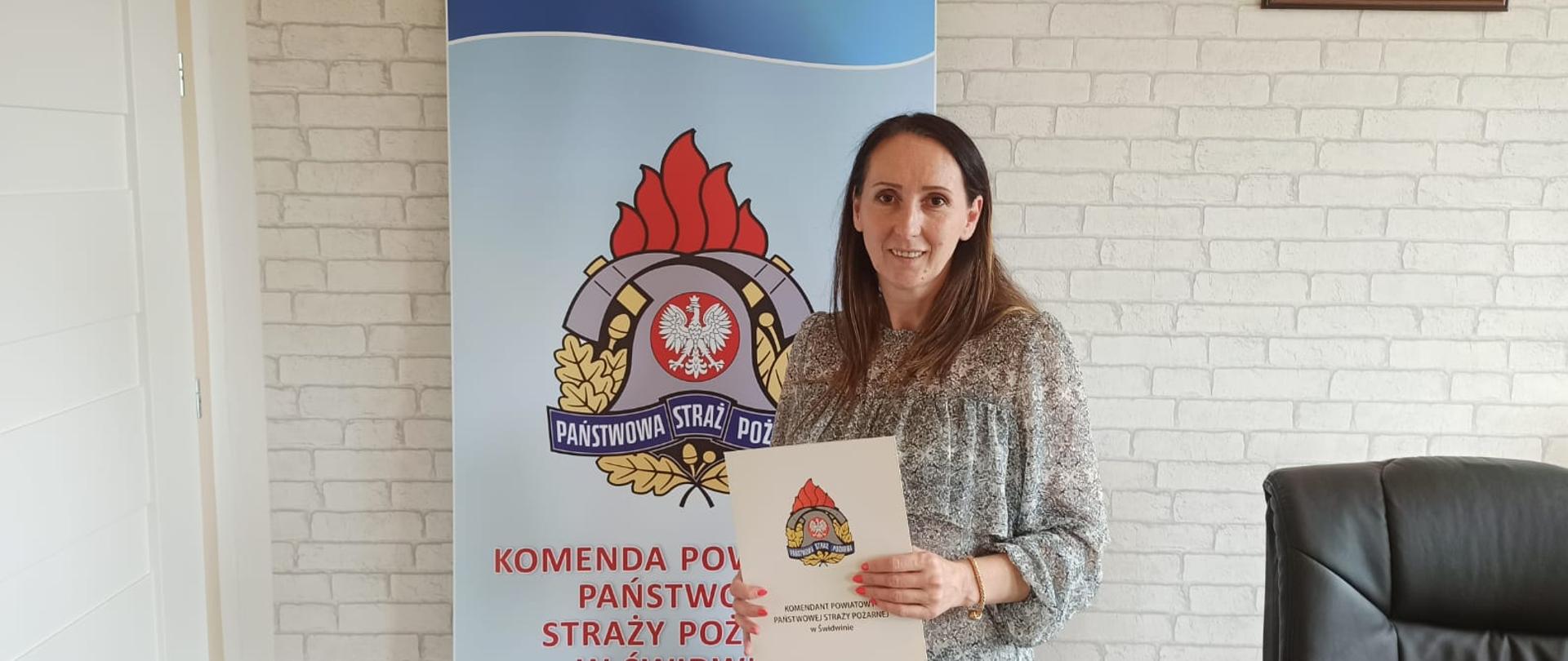 Zdjęcie przedstawia dwóch mężczyzn ubranych w czarne mundury strażackie oraz maseczki chirurgiczne, jeden z nich trzyma w rękach dokumenty. Za nimi widnieje baner z logiem PSP oraz napisem: "Komenda Powiatowa Państwowej Straży Pożarnej w Świdwinie". Za banerem widzimy ścianę z tapetą w szare cegły. Na ścianie wisi Godło Rzeczypospolitej Polskiej. Zdjęcie wykonano w gabinecie Komendanta Powiatowego PSP w Świdwinie.