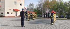 Na placu przed Jednostka Ratowniczo Gaśnicza nr 2 stoją strażacy w dwóch szpalerach naprzeciw siebie na uroczystej zbiórce. Pomiędzy szpalerami stoi dowódca. Strażacy ubrani są w stroje bojowe. Za szpalerami widoczny jest maszt na który poczet flagowy zawiesza biało-czerwona flagę.