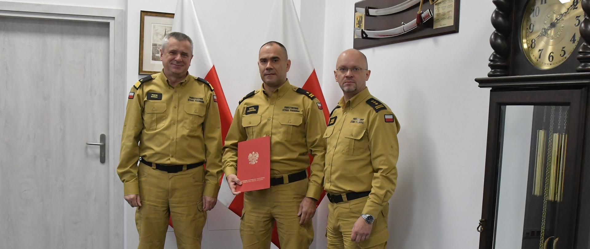 Trzech funkcjonariuszy Państwowej Straży Pożarnej stoi obok siebie strażak stojący w środku trzyma czerwoną teczkę za mężczyznami ustawione są flagi Polski na ścianie wisi obraz oraz szabla niedaleko ustawiony jest zegar. 