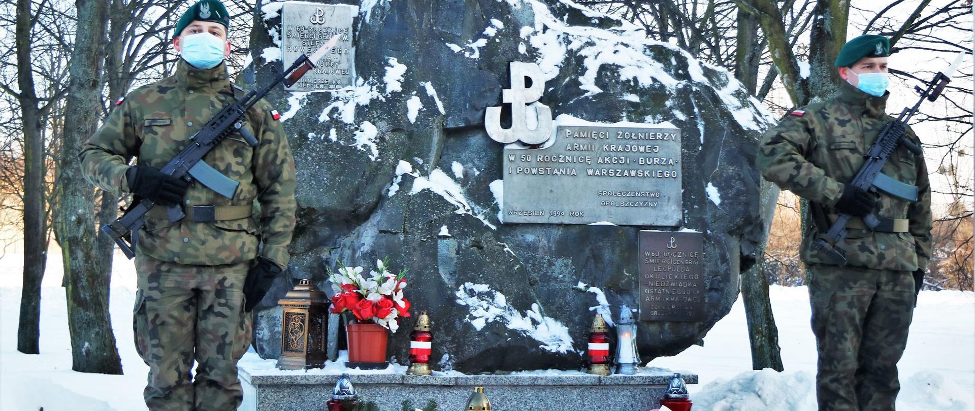 Na zdjęciu Pomnik Armii Krajowej w Opolu. Pomnik jest w centralnej pozycji kadru. To 2 metrowy postument w kształcie nieregularnego prostokąta. Po prawej jego stronie widać znak Polski Walczącej oraz napis Pamięci Żołnierzy Armii Krajowej. Po pomnikiem widać złożone kwiaty z biało-czerwonymi wstęgami oraz znicze. Po obu stronach pomnika stoją żołnierze, pełniący wartę honorową. W tle widać ośnieżone korony drzew. 