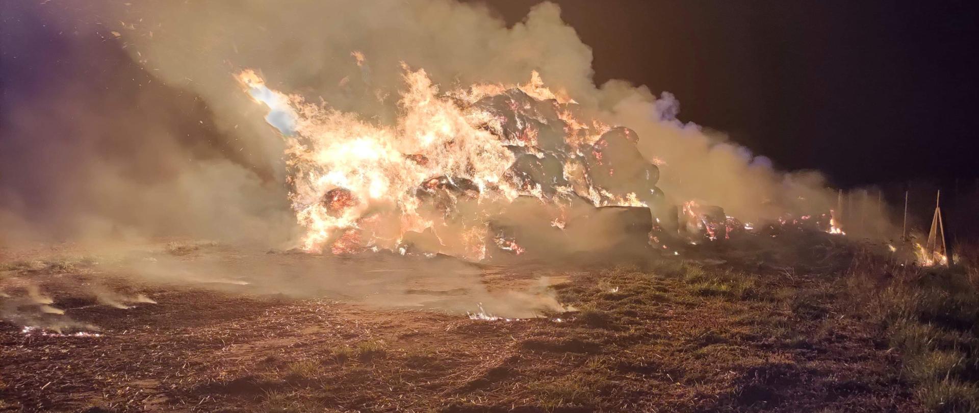 Zdjęcie przedstawia w całości objęty pożarem stóg siana, nad którym unosi się szary dym. Objęte pożarem baloty siana znajdują się na trawiastym podłożu, gdzie po prawej stronie zaobserwować można przebiegające ogrodzenie. Pożar ma miejsce w porze nocnej. 