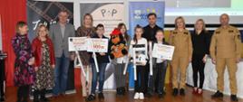 Grupa mężczyzn, kobiet i dzieci stoi w jednym szeregu, po prawej stronie widoczne osoby w mundurach straży pożarnej, stojące pośrodku dzieci trzymają dyplomy