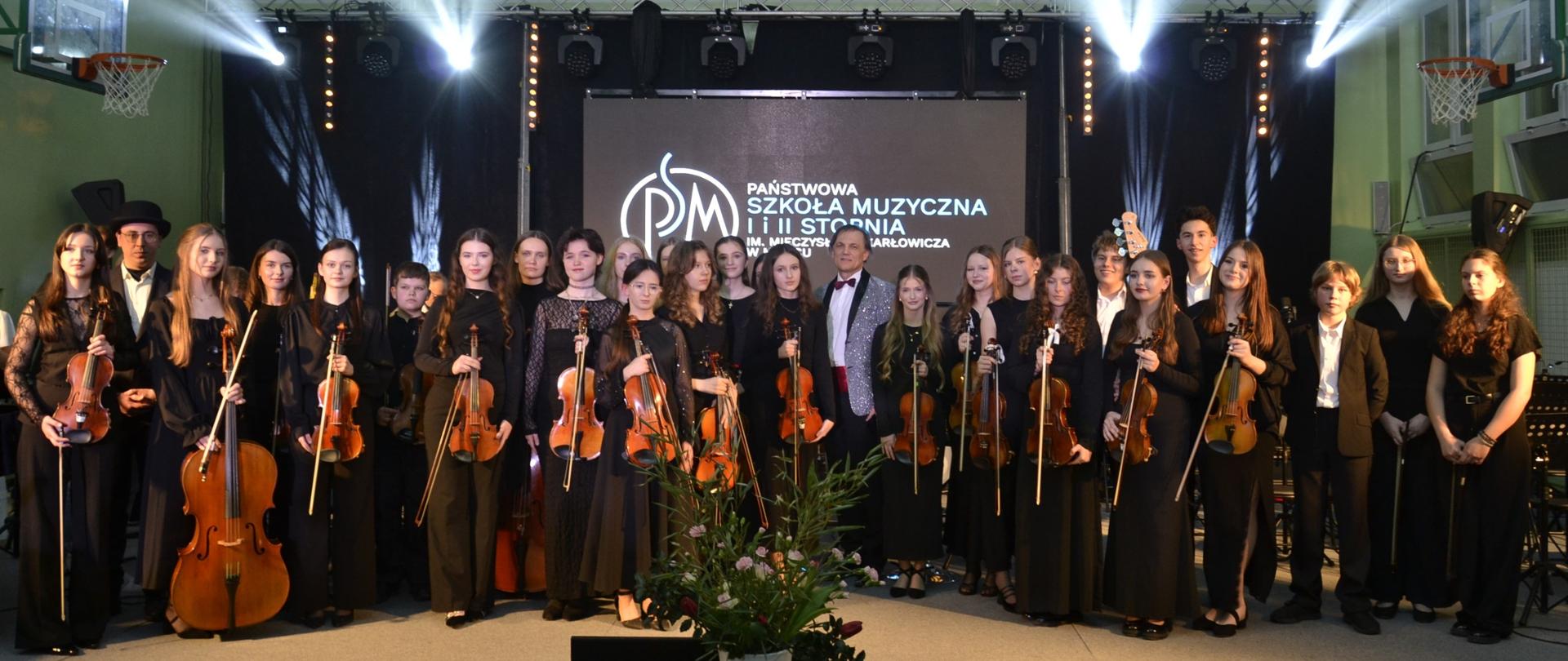 Członkowie Orkiestry Kameralnej i Orkiestry Dętej PSM II stopnia w Mielcu stoją na scenie po koncercie karnawałowym w sali I LO w Mielcu, na tle ekranu z logo szkoły muzycznej.