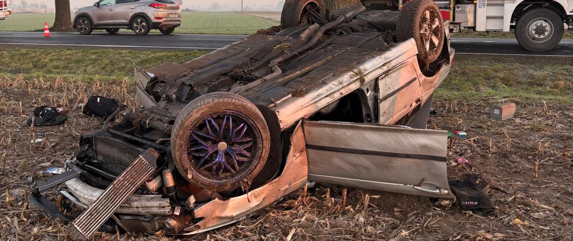 Srebrne auto osobowe leżąc na dachu pola uprawnego. W tle drzewo, wóz strażacki stojący na drodze.