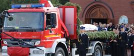 Samochód pożarniczy stoi przed kościołem na nim położona jest trumna obok stoi dwóch strażaków Państwowej Straży Pożarnej za nimi uczestnicy uroczystości.