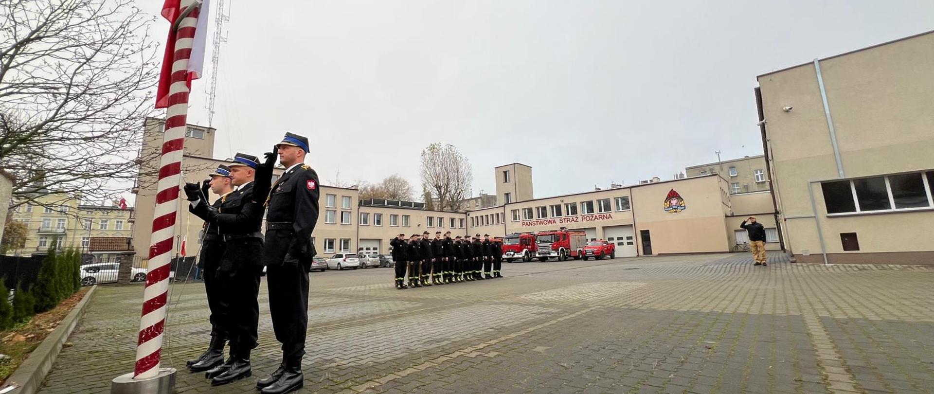 Uroczyste podniesienie flagi państwowej z okazji Święta Niepodległości - strażacy przy maszcie, w tle inni i strażnica