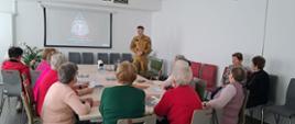 Na zdjęciu przedstawiono spotkanie informacyjno-edukacyjne z udziałem seniorów, odbywające się w jasnej, nowoczesnej sali konferencyjnej. W centralnej części pomieszczenia znajduje się prostokątny stół, przy którym zasiada kilkanaście osób – głównie kobiety w wieku senioralnym. Uczestnicy spotkania mają przed sobą materiały informacyjne i ulotki, a ich uwaga skierowana jest na funkcjonariusza Państwowej Straży Pożarnej, który prowadzi omówienie zagadnień związanych z bezpieczeństwem w domu i postępowania w przypadku zagrożenia. W tle widoczny jest ekran projekcyjny, na którym wyświetlone zostało logo Państwowej Straży Pożarnej – Komendy Powiatowej PSP w Wolsztynie. 