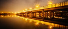 Nocna lub zmierzchowa scena przedstawiająca długi most rozciągający się nad spokojnym zbiornikiem wodnym. Most oświetlony jest szeregiem latarni ulicznych, których światło tworzy efekt gwiaździstych rozbłysków. Odbicia świateł i konstrukcji mostu w wodzie tworzą niemal symetryczny obraz. Niebo ma łagodny gradient – od ciemnego u góry do jaśniejszego przy horyzoncie. Na moście nie widać ludzi ani pojazdów.