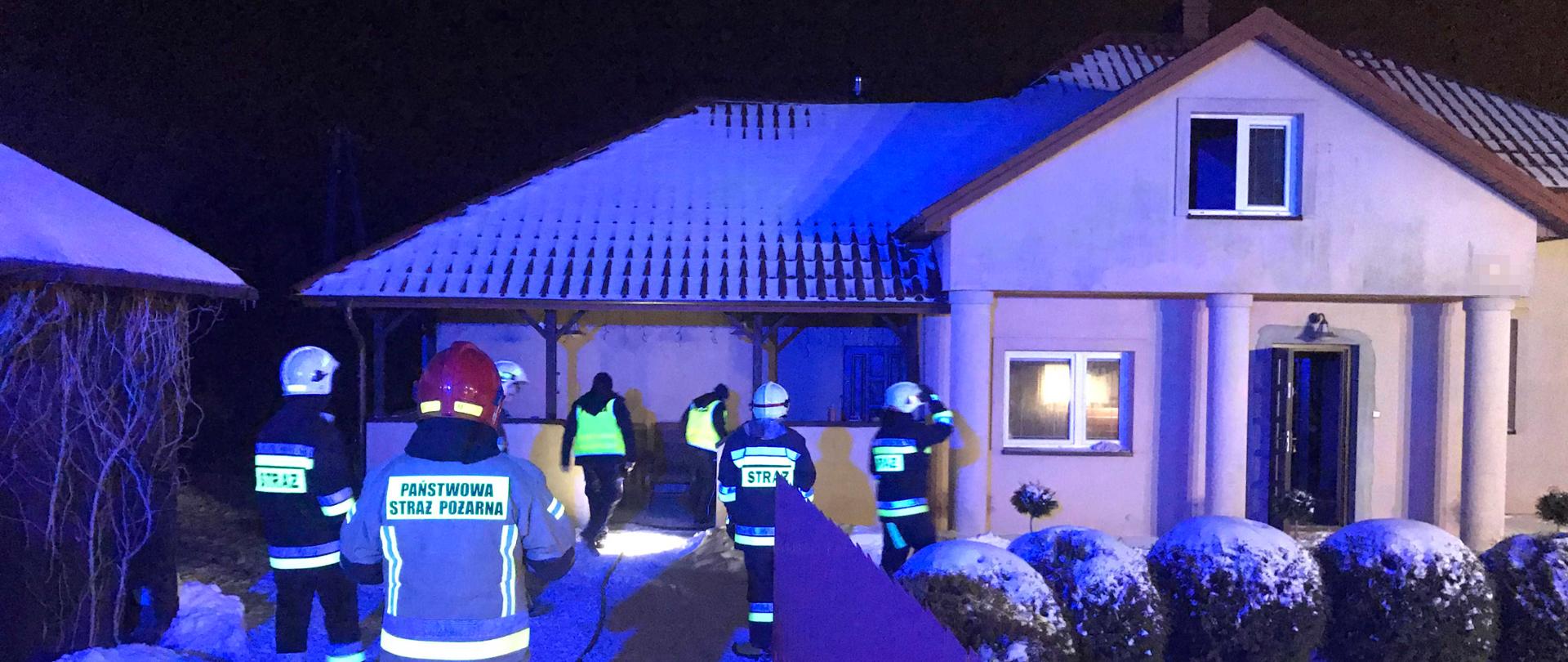 Alternatywny: zdjęcie zrobione w nocy. Zdjęcie przedstawia budynek mieszkalny, w którym pali się pomieszczenie kuchni. Obok domu stoi na ośnieżonym podjeździe pięciu strażaków i dwóch policjantów. 