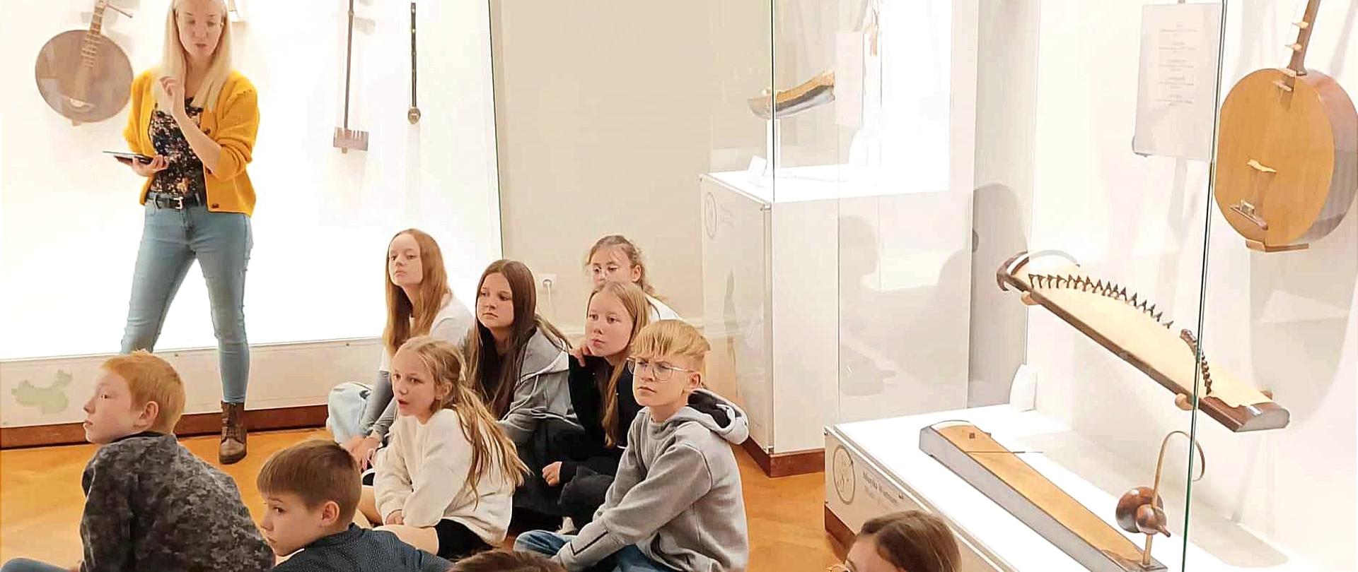Zdjęcie zrobione w Muzeum instrumentów Detych w Poznaniu. W sali na drewnianej podłodze siedza uczniowie 7 dziewczynek oraz 3 chłopców. W tle w gablotach znajduja się stare instrumenty muzyczne, które omawia pani z muzeum. 