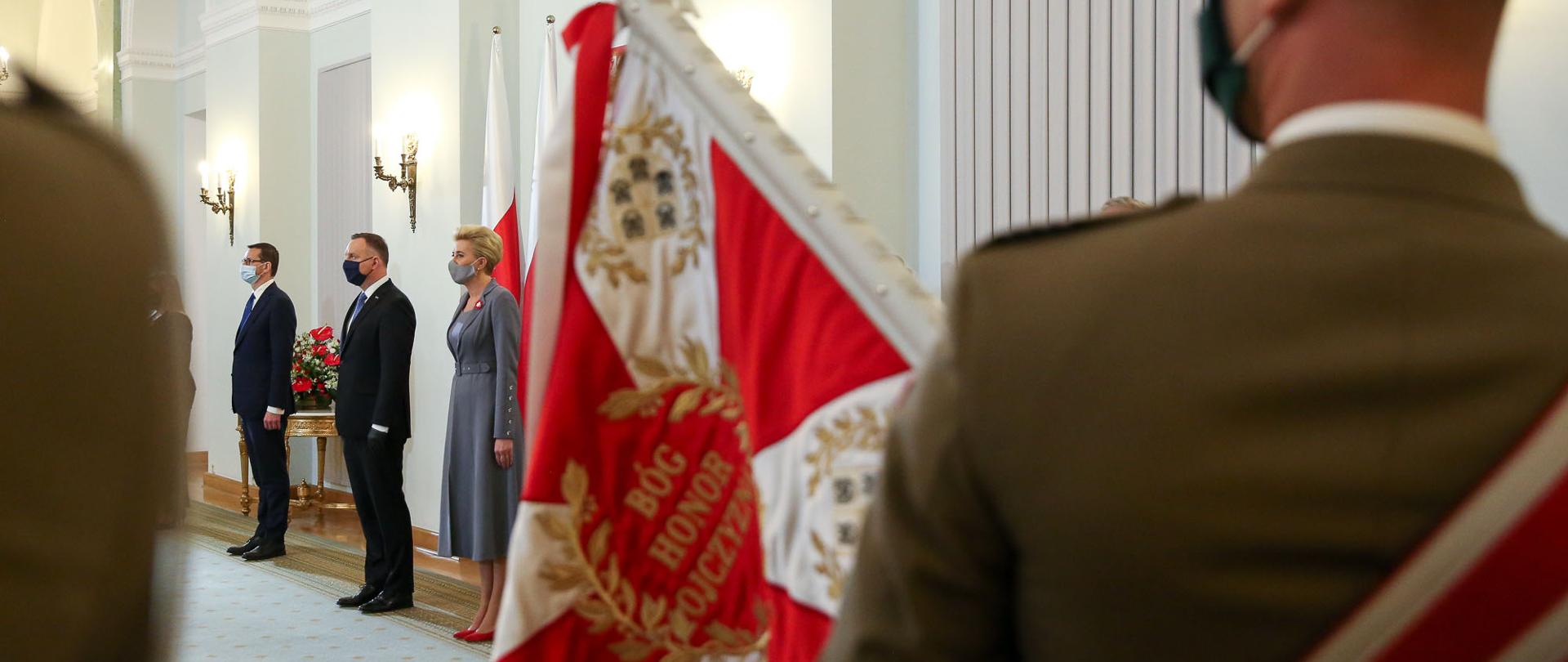 Widok zza sztandaru na stojących prezydenta Andrzeja Dudę, Agatę Dudę i premiera Mateusza Morawieckiego.
