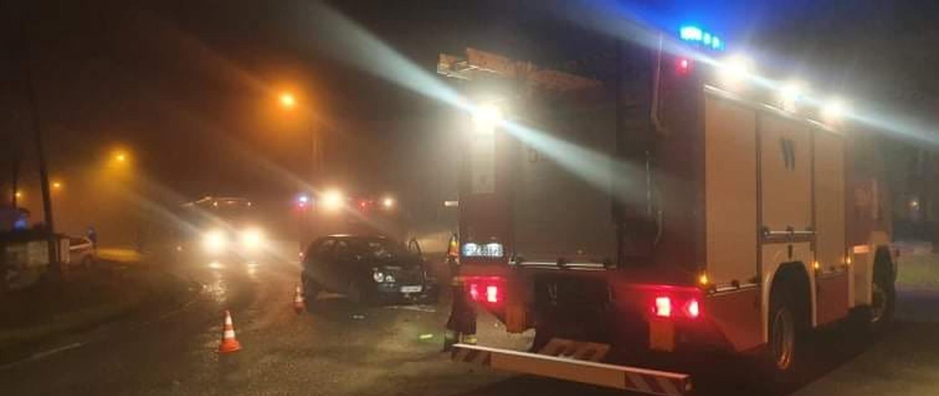 Widać dwa samochody strażacki, między nimi jeden uszkodzony samochód osobowy, jest ciemno