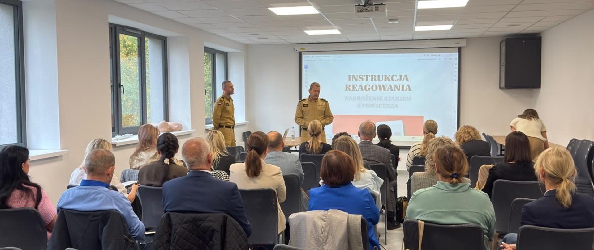 Komendant Powiatowy PSP w umundurowaniu służbowym koloru żółtego rozpoczyna spotkanie. Na przeciwko niego siedzą dyrektorzy szkół. 