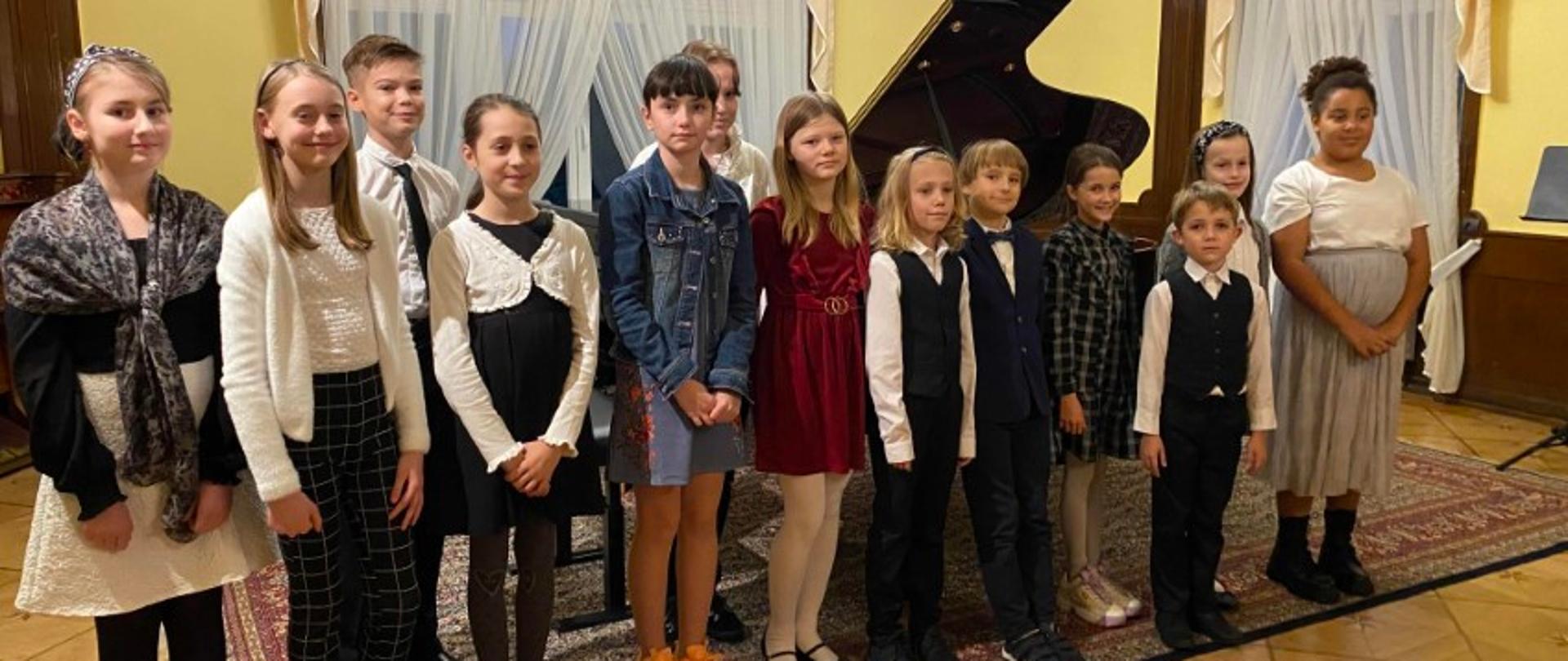 Zdjęcie kolorowe. Dziewczynki i chłopcy stoją na scenie, uśmiechają się, w tle widać fortepian.