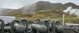 Zdjęcie przedstawia działające ciepłownicze instalacje geotermalne przy parkingu samochodowym w terenie górzystym w Islandii