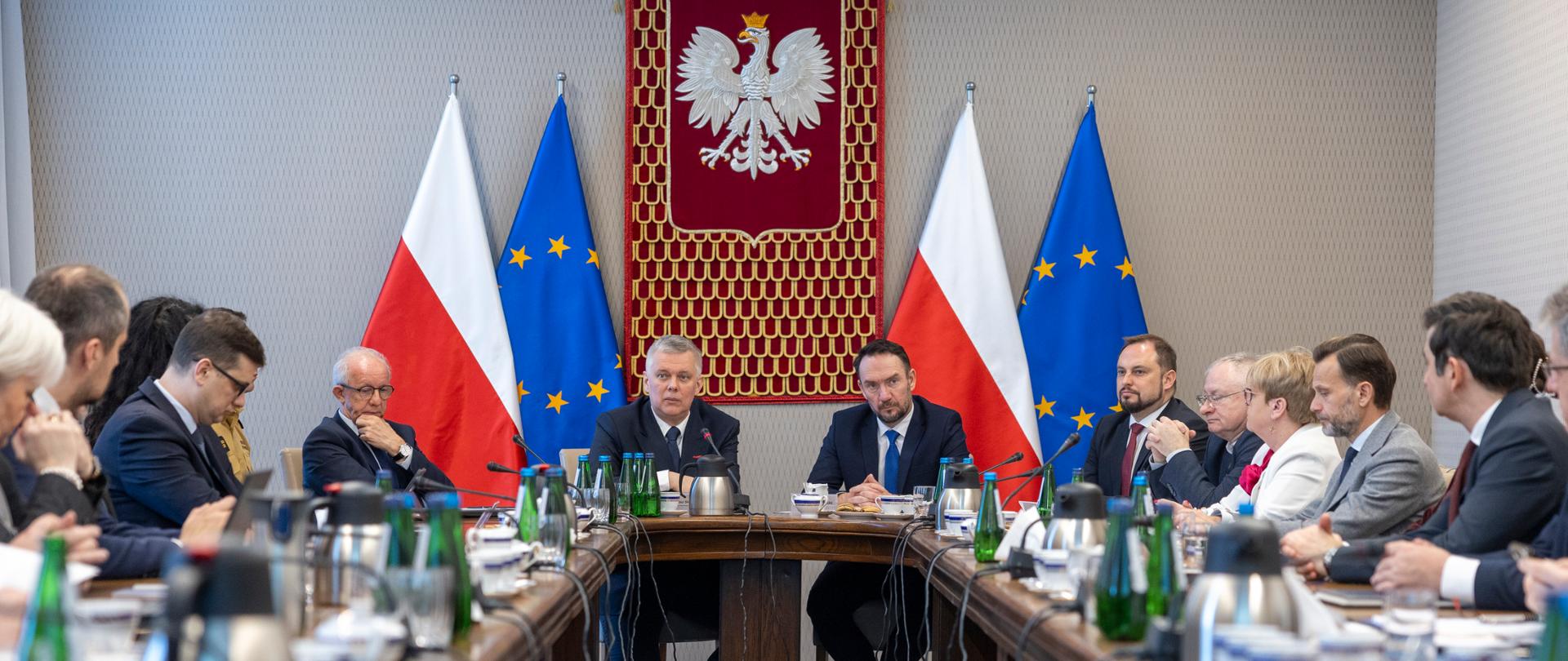 Spotkanie w MSWiA przy długim stole, z udziałem urzędników w formalnych strojach. W tle godło Polski oraz flagi Polski i UE.
