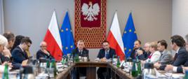 Spotkanie w MSWiA przy długim stole, z udziałem urzędników w formalnych strojach. W tle godło Polski oraz flagi Polski i UE.