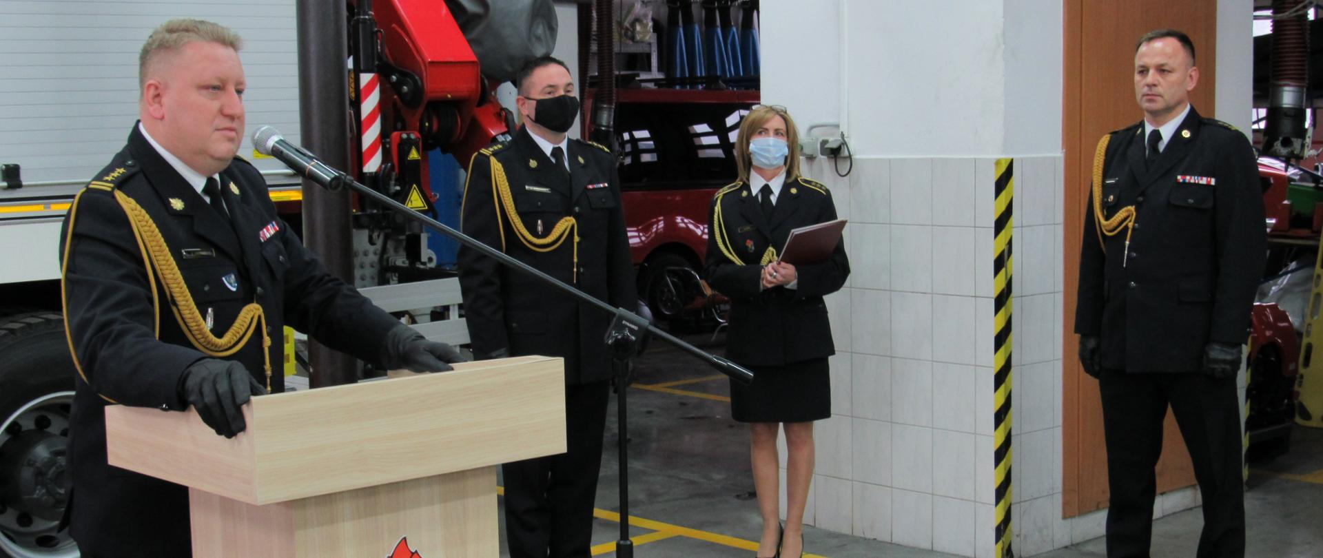 Zdjęcie przedstawia Łódzkiego Komendanta Wojewódzkiego PSP przemawiąjącego w trakcie zdaje w trakcie uroczystości zdania obowiązków komendanta powiatowego PSP w Zduńskiej Woli. Zdjęcie wykonano w garażu jednostki ratowniczo-gaśniczej w tle samochód gaśniczych, zdający obowiązki komendant powiatowy oraz dwoje strażaków. Wszyscy ubrani w umundurowanie wyjściowe.