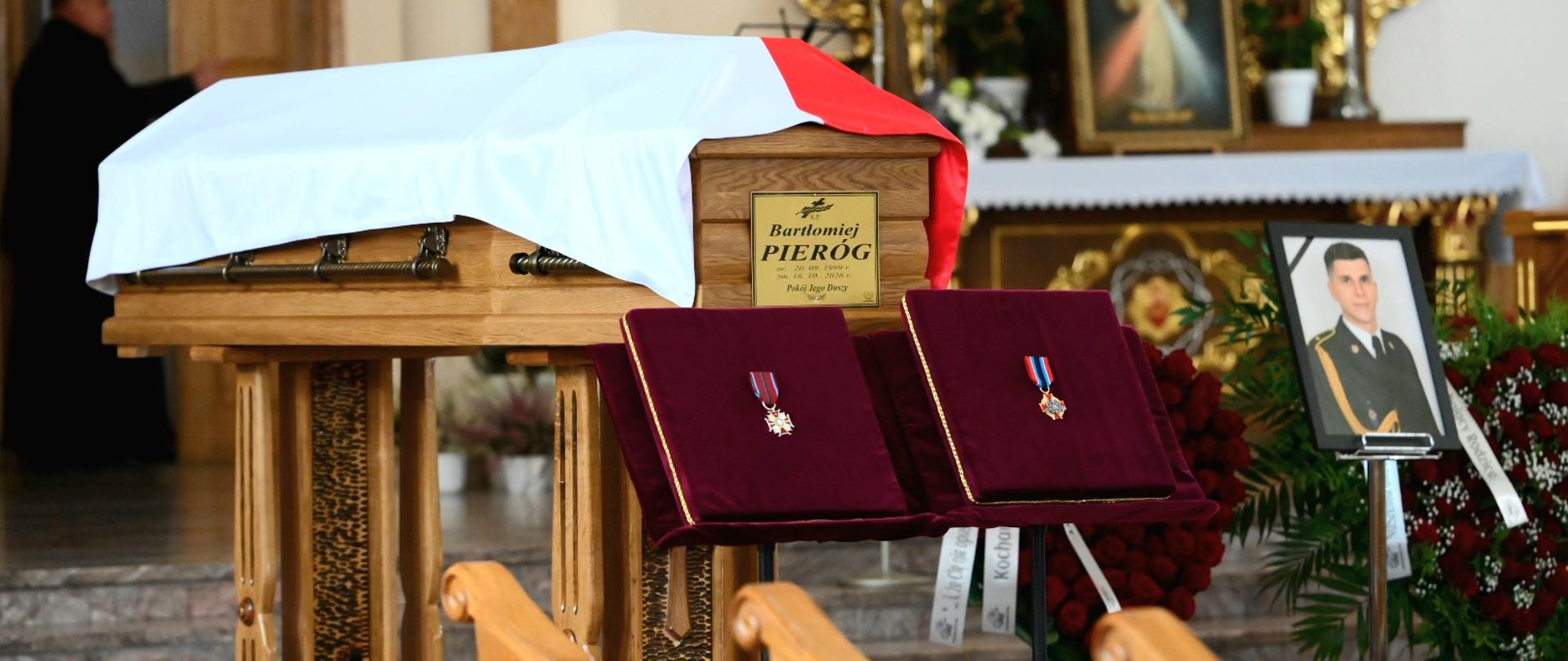 Zdjęcie 3: Trumna Bartłomieja Pieroga przykryta biało-czerwoną flagą ustawiona przed ołtarzem w kościele. Przed nią znajdują się odznaczenia, a obok portret zmarłego strażaka i wieńce pogrzebowe.
