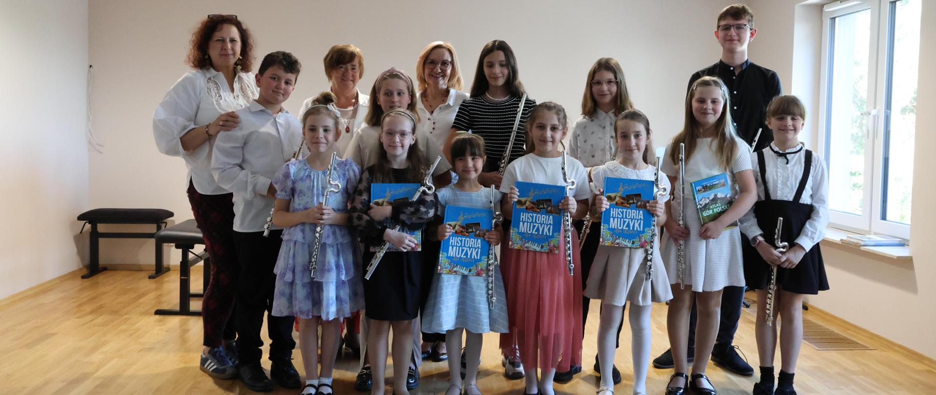 Grupa uczniów z nauczycielkami stoi na scenie sali koncertowej, każde dziecko trzyma flet i książkę