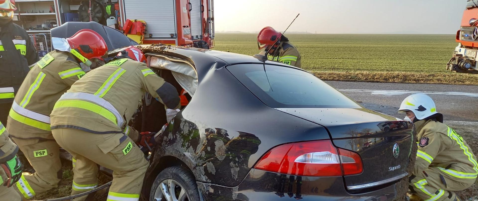 Obraz przedstawia rozbity samochód osobowy. Przy pojeździe znajdują się strażacy w umundurowaniu specjalnym wykonujący dostęp do poszkodowanego mężczyzny uwięzionego w pojeździe. W tle samochody pożarnicze.