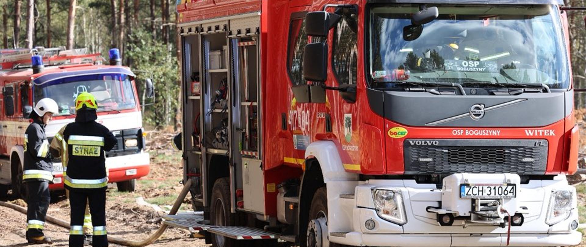 Na pierwszym planie widać czerwony samochód strażacki podczas akcji gaśniczej. Z lewej strony widać dwóch rozmawiających ze sobą strażaków w granatowych mundurach. W tle widoczny jest las.