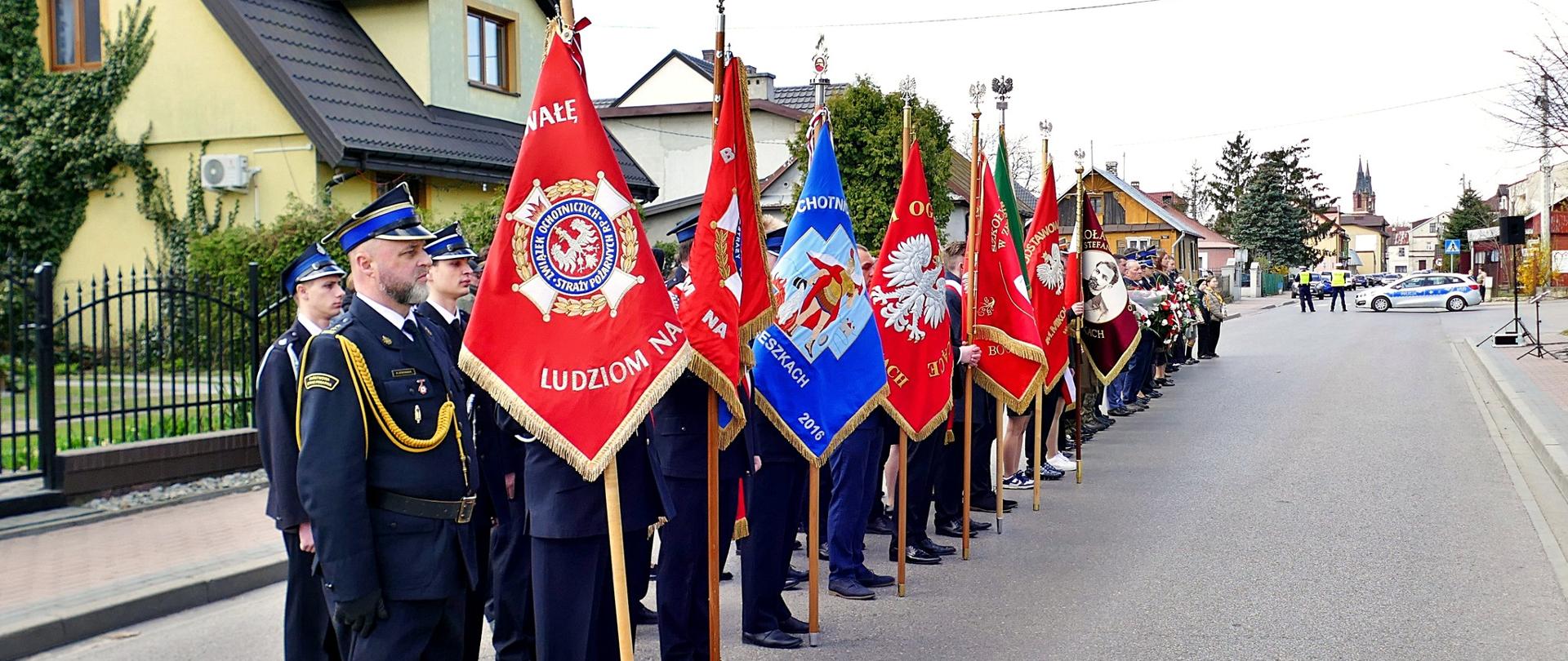 Poczty sztandarowe złożone z przedstawicieli służb mundurowych i organizacji lokalnych stoją w szeregu na ulicy podczas uroczystości patriotycznej w Łosicach.