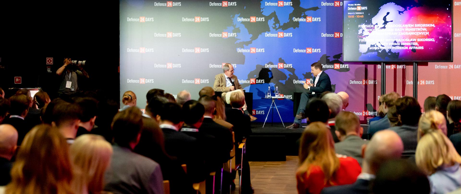 Na zdjęciu jest dwóch mężczyzn, którzy siedzą na scenie. Przed nimi jest kilkanaście osób na krzesłach. Są zwrócone w kierunku sceny. Z tyłu jest mapa. 