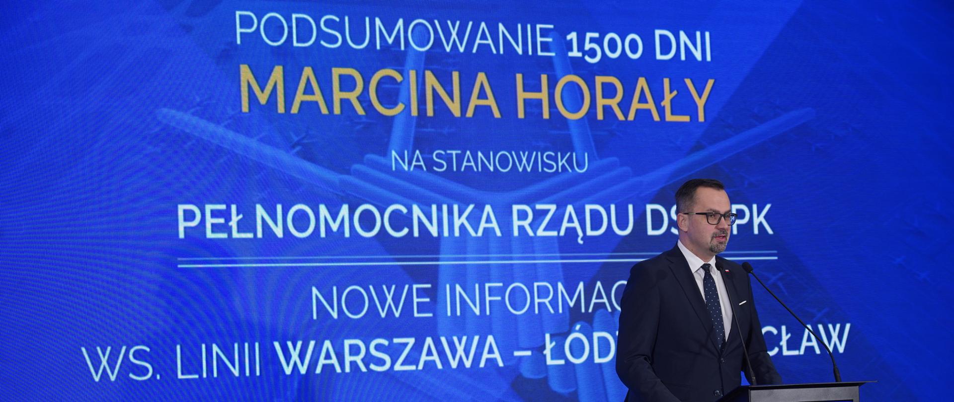 Wiceminister Marcin Horała stoi z prawej strony przy pulpicie i mówi do mikrofonu.