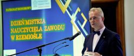 Za mównicą stoi mężczyzna w garniturze, mówi do mikrofonu, za nim niebieski ekran z napisem Dzień mistrza nauczyciela zawodu w rzemiośle.