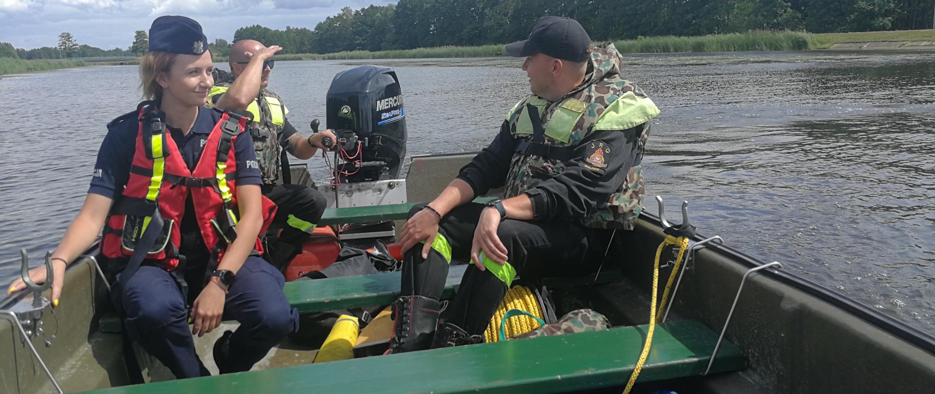 Zdjęcie przedstawia pokład łodzi na którym znajdują się strażacy oraz policjantka. Pasażerowie ubrani są w mundury charakterystyczne dla ich formacji oraz kamizelki asekuracyjne. Jeden ze strażaków siedzi za sterami silnika łodzi. Na podłodze widoczny jest różny sprzęt do ratownictwa wodnego. W tle widoczny jest zalesiony brzeg rzeki.